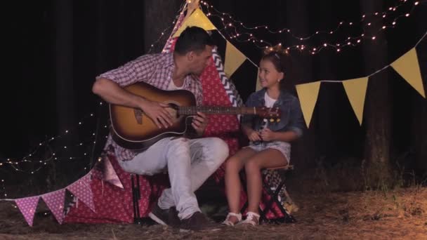 Relationship of relatives, rejoice male parent with his female child playing guitar having fun in the woods on background of wigwam during night picnic — ストック動画