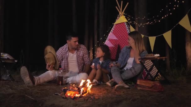Krewni na pikniku, radosna dziewczynka z rodzicami słuchają wesoło zabawnych opowieści i piją sok przy ognisku na tle wigwam podczas kempingu w lesie — Wideo stockowe