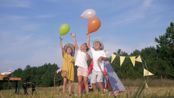 Cheerful childhood, friends boy and girls having fun at childly picnic jump and release balls on forest meadow against background of wigwam — Stock Video