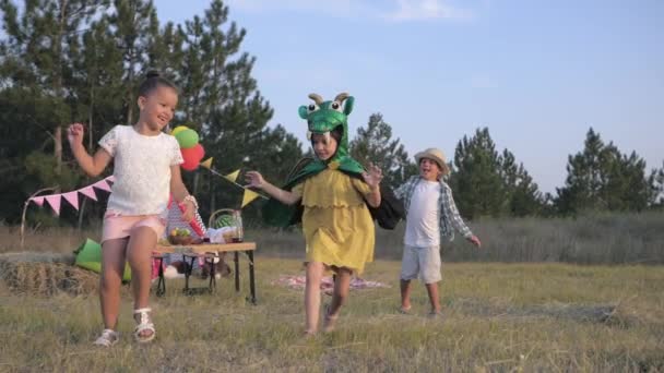 재미있는 아이들, 카니발 의상을 입은 어린 소녀는 친구들과 함께 게임을 하고 자연에서 피크닉을하는 동안 wigwam의 배경에 숲 의 숲 글레이드에서 그들을 따라 실행합니다. — 비디오