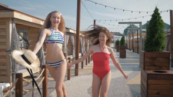 Fröhliche Freundinnen mit schlanken Körpern in Badeanzügen laufen Händchen haltend während des Sommerurlaubs im luxuriösen Resort — Stockvideo