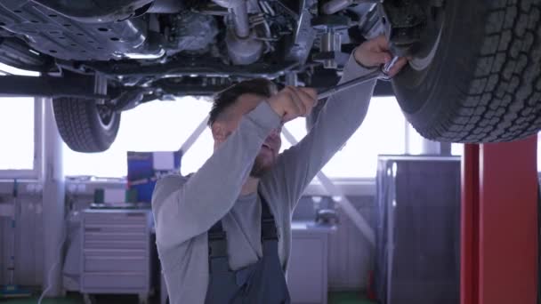 Arbeit in der Werkstatt, gut aussehender Mechaniker mit Schraubenschlüssel arbeitet unter dem Boden des Autos in der Werkstatt — Stockvideo