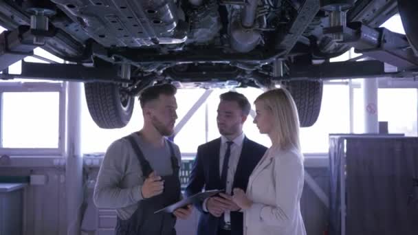 Mitarbeiter des Autoservice beraten Frau und Mann mit Klemmbrett in den Händen, die unter einem auf einem Hydrauliklift angehobenen Auto stehen — Stockvideo