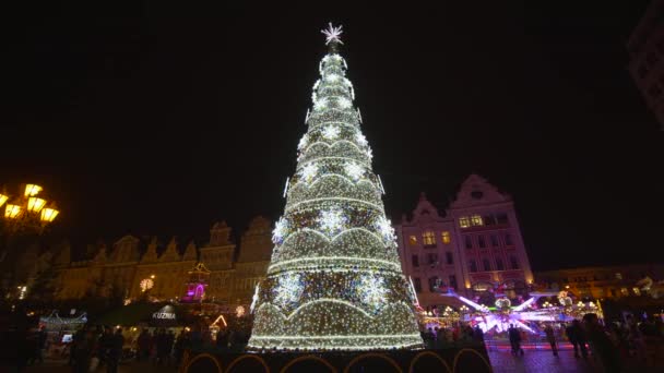 Туристы прогуливаются возле новогодней елки с блестящими гирляндами и красочными игрушками на Рыночной площади во время отпуска вечером — стоковое видео