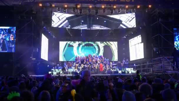 Толпа людей на концертном стенде возле сцены с мигающими прожекторами на открытом воздухе в вечернее время — стоковое видео