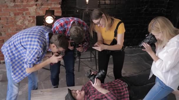 Cameramans com equipamento profissional no fundo de softboxes e parede de tijolo tirar fotos menina de chapéu que está deitado em uma mesa com câmera — Vídeo de Stock