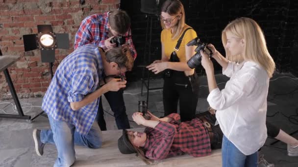 Sesión de fotos en el estudio, cámaras con equipo profesional en el fondo de softboxes y pared de ladrillo tomar fotos una chica alegre en el sombrero que está acostado en la mesa con una cámara — Vídeos de Stock