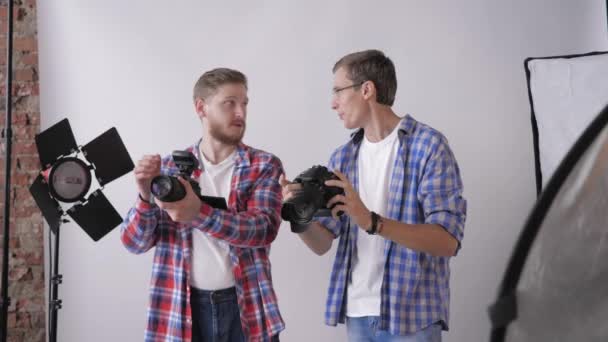 Fotógrafos creativos en cámaras digitales en manos discuten sesión de moda en estudio fotográfico durante taller de fotografía sobre fondo de lámparas de iluminación — Vídeo de stock