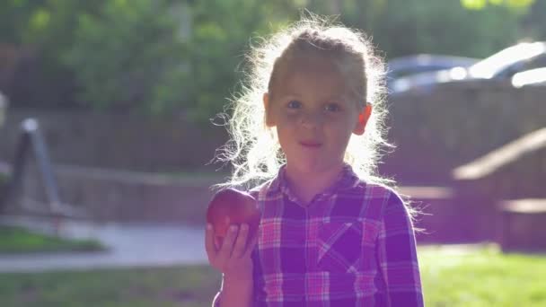 Kind bijt appel in achtergrondlicht, meisje gekleed shirt kauwt sappig rijp fruit en kijk op camera naar de natuur — Stockvideo