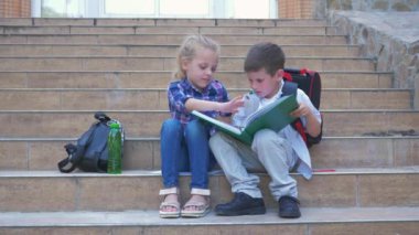Sırt çantalı öğrenciler açık havada okulun merdivenlerinde oturmuş kitap okuyor ve sayfaları karıştırıyorlar.