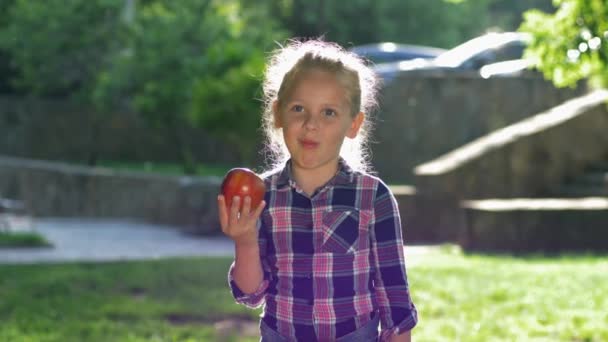 Счастливая маленькая блондинка укусы и смешно жевать спелые сочные яблоко в подсветке, ребенок одет рубашку смотреть на камеру и улыбается на открытом воздухе — стоковое видео