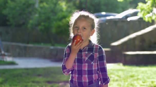 Porträt eines essenden Kindes im Gegenlicht, kleines blondes Mädchen kaut reifen, saftigen Apfel und blickt im Freien in die Kamera — Stockvideo