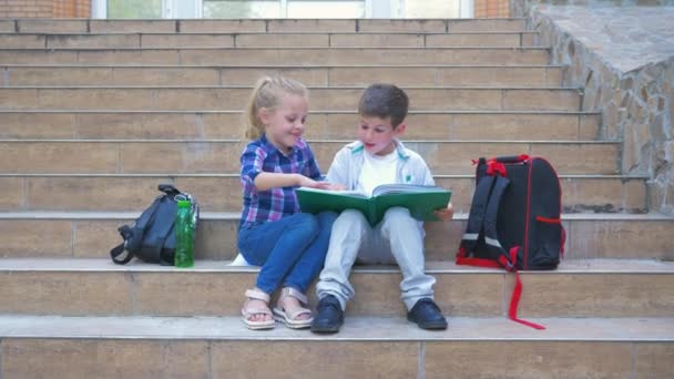Escuela primaria, alumnos con mochilas sentados en los escalones y hojeando un libro durante el recreo al aire libre — Vídeos de Stock