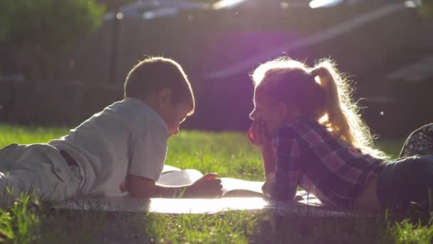 Eerste liefde, mooi jongetje en meisje liggend op gras en foto 's bekijken in een boek tijdens schoolvakanties in het zonlicht — Stockvideo