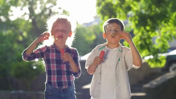 Feliz infancia, niña y niño soplando burbujas de jabón en el parque jugando al aire libre en la luz de fondo — Vídeo de stock
