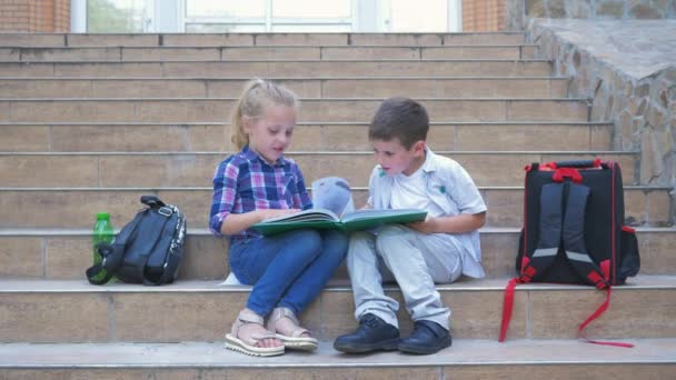 Маленькие одноклассники с рюкзаками сидят на ступеньках школы и просматривают книгу во время перерыва на свежем воздухе — стоковое видео