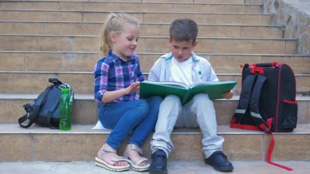 Pequeños estudiantes de la escuela con mochilas sentadas en los escalones y hojeando un libro durante el recreo al aire libre — Vídeos de Stock