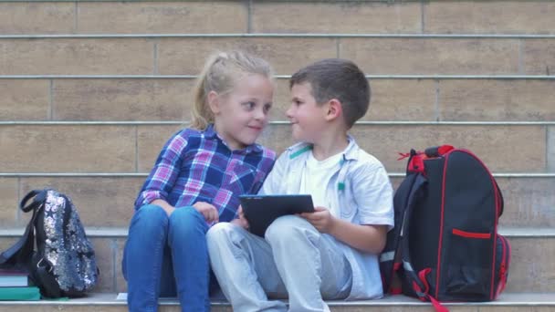 Pequeños alumnos modernos utilizan gadget sentado en los escalones de la escuela con mochilas durante el recreo al aire libre — Vídeos de Stock