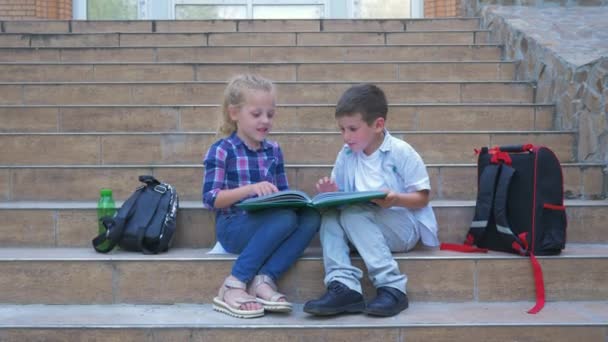 Descanso escolar, niño y niña se comunican y revisan libro sentado en los pasos de la escuela junto a las mochilas al aire libre — Vídeo de stock