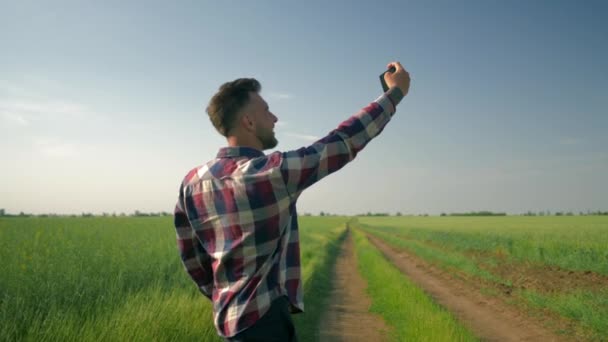 Cheerful farmer in checkered shirt hold cell phone and takes selfie photo on background of green field and blue sky — ストック動画