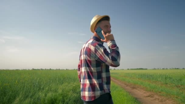 Agriculteur parlant au téléphone tout en marchant sur une route de campagne, homme souriant au chapeau et chemise à carreaux profiter de l'air frais et la beauté de la nature sur vert — Video