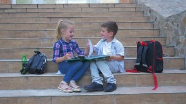 Okul çocukları okul merdivenlerinde sırt çantalarının yanında oturuyorlar ve açık havada kitap sayfalarını karıştırıyorlar.