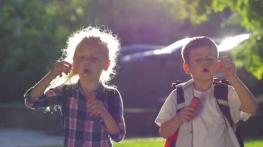 Okul çocukları. Sırt çantalı bir kız ve erkek. Açık havada arka planda bir dizi sabun köpüğü üflüyorlar.