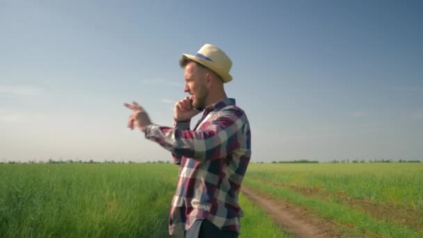 Joyful farmer in straw hat and checkered shirt talking on a cell phone with happy emotions during walking along country road in his field — Stock Video