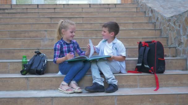Schoolkinderen die op school naast rugzakken zitten en tijdens de pauze in de open lucht door het boek bladeren — Stockvideo