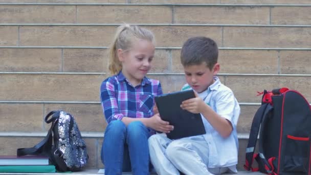Okul çocukları tatilde sırt çantalarıyla okul merdivenlerinde otururken dijital tablet kullanıyorlar. — Stok video