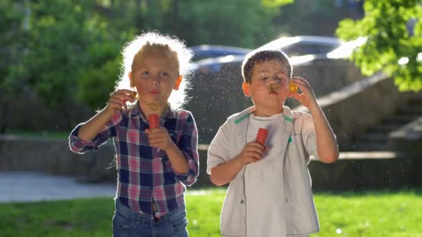 Two little children boy and girl playing outdoors and blowing soap bubbles in backlight close up — Stock Video