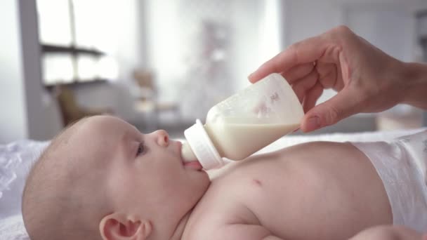 Portrait of baby girl drinking milk from a bottle that mother holds in her hands, newborn in diaper is lying on changing table close-up — Stock Video