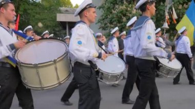 Yürüyüş bandosu, beyaz üniformalı genç denizciler büyük davullarla çalıyorlar ve Ukrayna bayrağını yavaş çekimde sokakta taşıyorlar.