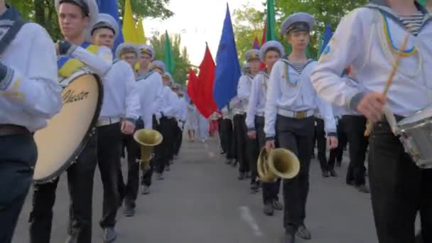 Молодые моряки в форме с палками в руках играют на больших барабанах во время марша и несут красочные флаги на улице, девушка в параде машет руками перед камерой — стоковое видео