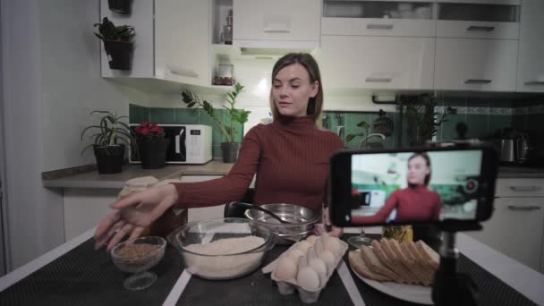 Güzel kadın blogcu mutfakta erzakla oturmuş abonesi için akıllı telefona video tarifi yazıyor sonra tost yaparken bir bardak şarap ve içecek kaldırıyor. — Stok video