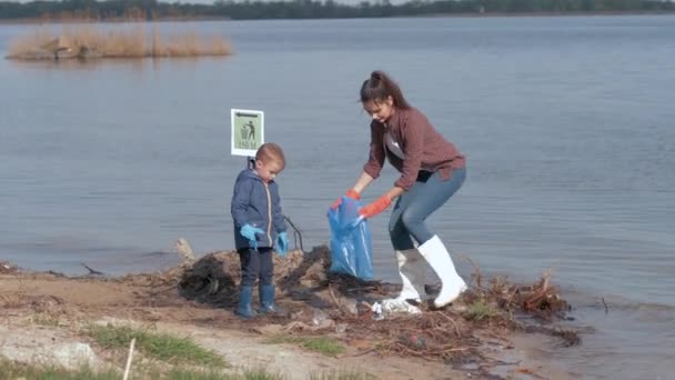 Zorg natuur ecologie, kind jongen helpt vrouwelijke vrijwilliger activist opruimen vervuilde rivier kust van plastic afval en verzameld in vuilniszak redden milieu — Stockvideo