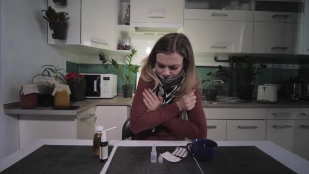 Симптомы заболевания, женщина чувствует себя больной кашляет и замерзает из-за температуры, сидя за столом с лекарствами и пьет горячий напиток от боли в горле — стоковое видео