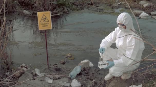 Bio-Gefahr in der Natur, Gefahrstoffchemiker in Schutzanzug entnimmt infizierte Wasserprobe in Reagenzgläsern für Tests in kontaminiertem Fluss mit Zeiger auf biologische Gefahr — Stockvideo