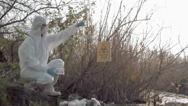 Биологическая опасность в природе, исследователь химзащиты в защитном костюме взяв образец зараженной воды в пробирках для тестирования в загрязненном заливе с указателем bi — стоковое видео