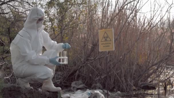 Farligt arbete, hazmatbiolog i skyddskläder som tar smittat vattenprov i provrör för undersökning i förorenad loch med pekartecken biologisk fara — Stockvideo