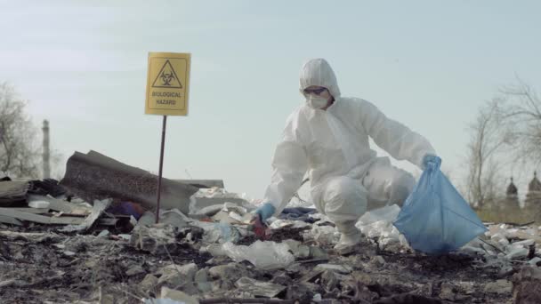 Umweltverschmutzung: Gefahrstoffermittler in Schutzanzug sammelt Abfall im Müllsack für Forschung auf Müllhalde mit Hinweisschild Biologische Gefahr — Stockvideo