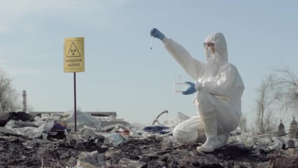 Peligro de radiación, virólogo Hazmat en uniforme tomar la muestra de basura infectada en el tubo de ensayo para examinar en el vertedero de basura con signo de puntero peligro de radiación — Vídeos de Stock