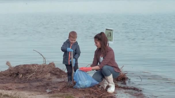 Ekologická péče, maminka s chlapečkem dobrovolníci čistí znečištěnou přírodu z plastového odpadu u vody na nábřeží — Stock video