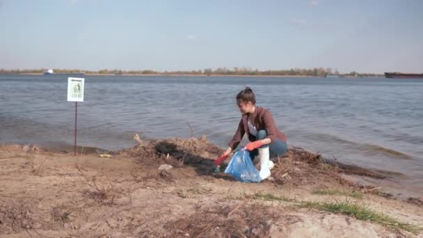 Ecologisch probleem oplossen, jonge vrouw vrijwilliger reinigt vuil strand en verzamelt plastic afval in vuilniszak en geeft duim omhoog op de rivier — Stockvideo
