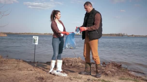 Екологічна природа, пара волонтерів-активістів очищають забруднену набережну річки від пластикових сміття і збираються в сміттєвому мішку, рятуючи екологічну природу — стокове відео