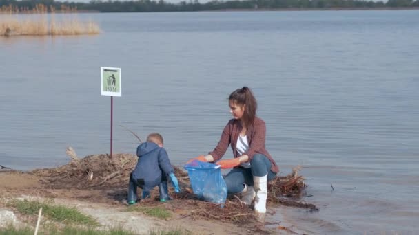 환경 문제 해결을 위해 소년은 여성 자원 봉사 활동을 하는 여성을 도와 플라스틱 쓰레기에서 더러운 강을 정화하고 생태학을 보존하기 위해 쓰레기 봉투에 모아 진다 — 비디오