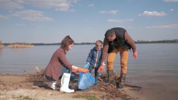 Protección del medio ambiente, la mujer y el hombre voluntarios con niño recogen plástico y polietileno basura en terraplén sucio cerca del agua — Vídeo de stock