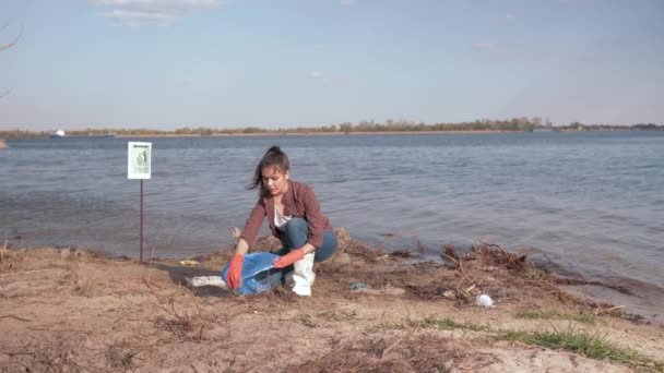 Soluciones ambientales, chica voluntaria recoge basura de plástico en la bolsa de basura y da gesto positivo en la playa del río contaminado cerca del agua — Vídeo de stock