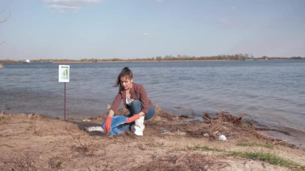 Environmental solutions, girl volunteer collects plastic trash in garbage bag and gives positive gesture on polluted river beach near water — Stock Video