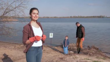 Kirliliğe karşı genç toplum, gülümseyen kadın gönüllü portresi, plastik çöpten plajı temizlerken küçük çocuklu ailenin odaklanmamış geçmişine parmak basıyor.
